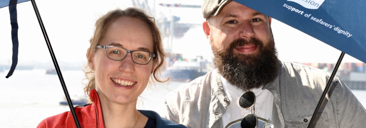Die Seemannsmission sucht Partner. Für unsere Arbeit brauchen wir Partner sowohl für einmalige und langfristige Projekt-Kooperationen. Sprechen Sie uns gerne an. Das Foto zeigt Stefanie Langos (li.) und Diakon Fiete Sturm. Beide halten Schirme mit dem Logo der Deutschen Seemannsmission vor dem Hafenpanorama an der Elbe in die Kamera. Es ist Sommer, die Sonne scheint.