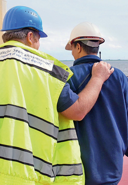 Von hinten gesehen: ein Mitarbeiter der Seemannsmission mit Leuchtweste steht neben einem Seemann in blauem Overall und legt ihm die Hand auf die Schulter- Symbolbild PSNV