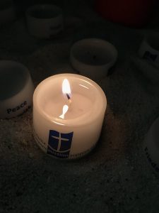 Brennende kleine weiße Kerze mit den Logo der Deutsche Seemannsmission, einer blauen Flagge mit einem weißen Anker Daneben eine Kerze auf der Peace steht