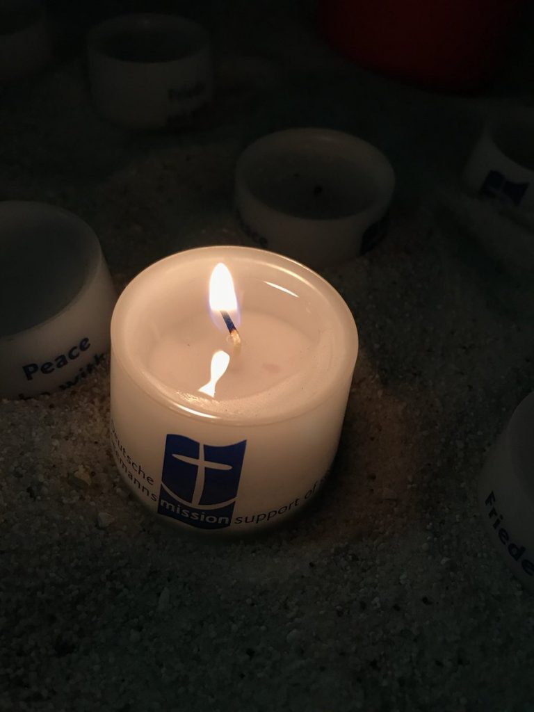 Brennende kleine weiße Kerze mit den Logo der Deutsche Seemannsmission, einer blauen Flagge mit eienm weißen Anker Daneben eine Kerze auf der Peace steht
