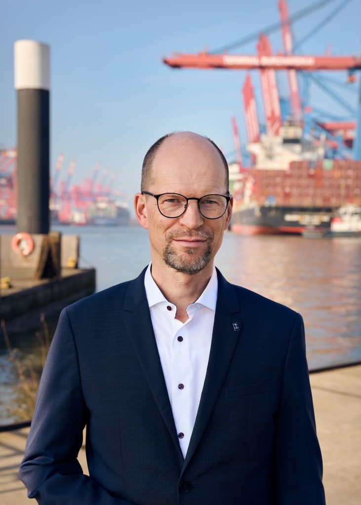 Generalsekretär Matthias Ristau steht in dunkelblauem Sakko und weißem Hemd vor dem Hafen. Liecht verschwommen sind Containerschiffe zu sehen.