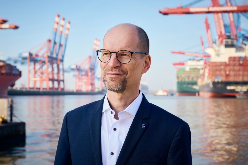 Matthias Ristau, Generalsekretär der Deutschen Seemannsmission steht mit dunkelblauem Sakko und weißem Hemd vor dem Containerhafen, er blickt leicht nach links
