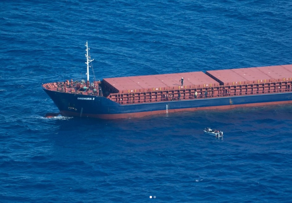 Die Luftaufnahme zeigt die vordere Hälfte eiens Frachtschiffs mit blauem Rumpf und rotem Deck. Eine PErson steht an Bord, Rundum ist Wasser und ein kleiens Boot mit ca. 20 Personen 
