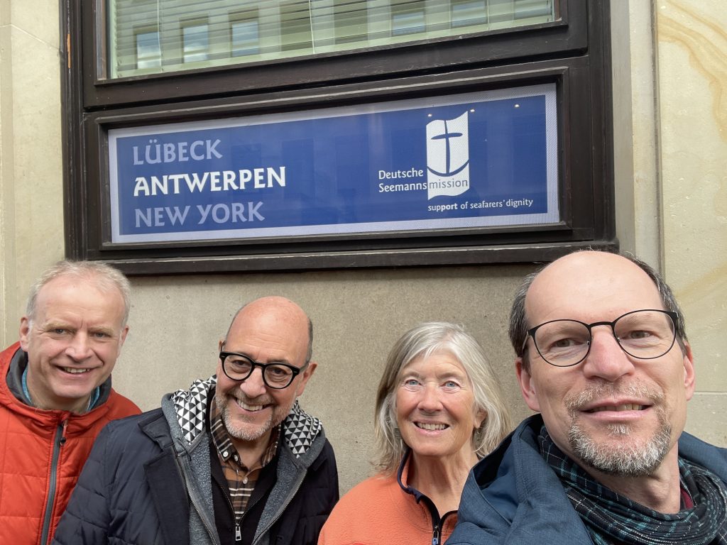 eine Frau und zwei Männer stehen vor einem Fenster auf dem steht: Lübeck, Antwerpen, New York. 
