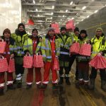 Acht Seemänner stehen im großen Innendeck der Bore Sea, eines RoRo Schiffs. Sie tragen gelb leuchtende Arbeitskleidung und halten rote Tüten mit den Geschenken in der Hand - Weihnachtsglück an Bord