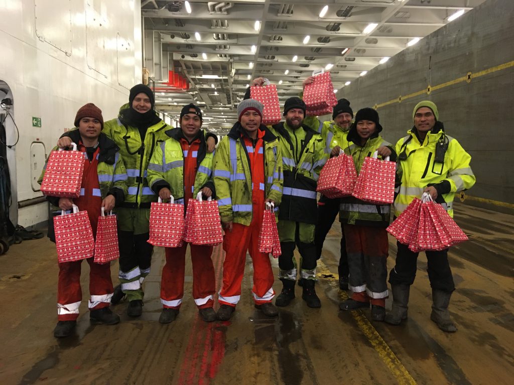 Acht Seemänner stehen im großen Laderaum der Bore Sea, eines RoRo Schiffs. Sie tragen leuchtend gelbe Arbeitsjacken und halten rote Tüten mit Geschenken in der Hand - Weihnachtsglück an Bord. 