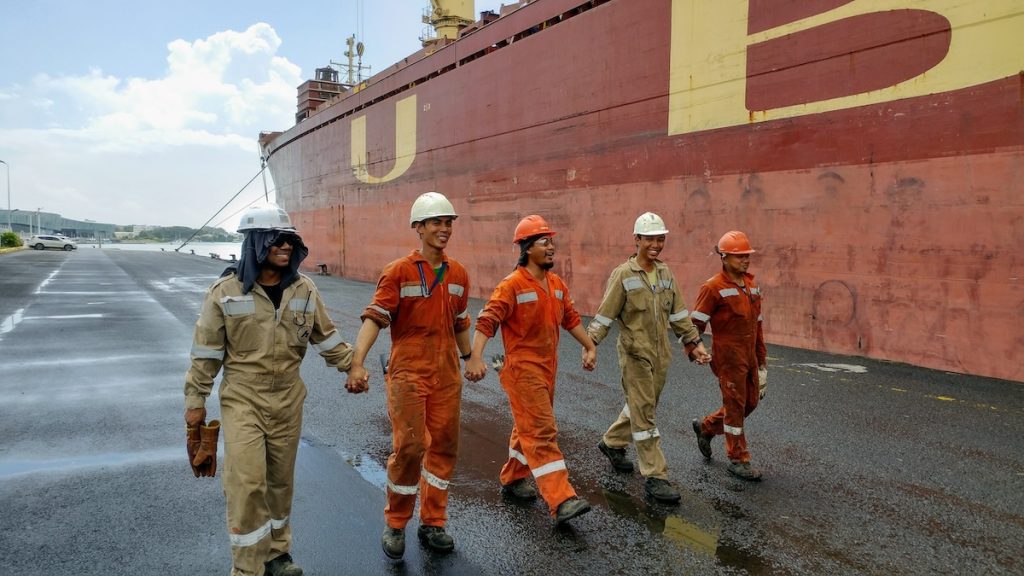 Fünf Seemänner gehen in einer Kette, sich die Hände gebend auf dem Kai neben einem großen roten Schiff - Fotos von Seeleuten - Ausstellung vom ITF Seafarers Trust