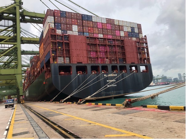 Zu sehen ist ein schwarzes Containerschiff, das viele bunte Container geladen hat. Es ist die Al Jasrah, die an den Kaianlagen im Hafen von Singapur liegt. 
Die Seemannsmission hat die Seeleute betreut. Sie sagt: "Rotes Meer für Seeleute lebensgefährlich."