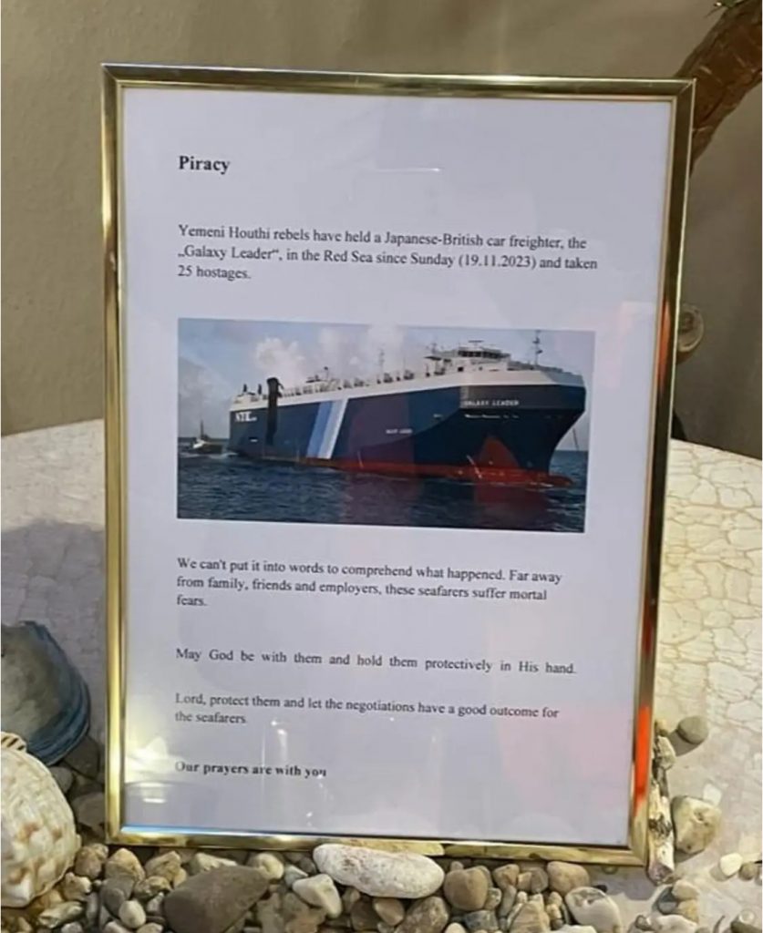 Auf ein paar Kieselstein steht ein Bilderrahmen mit goldenem Rahmen. Drinnen ist ein weißes Blatt mit Text. Das WOrt Piracy, englisch für Piraterie ist zu erkennen. Und ein Foto des blauen großen Schiffs, der Galaxy Leader. Seit 3 Monaten sind die Seeleute als Geiseln festgehalten