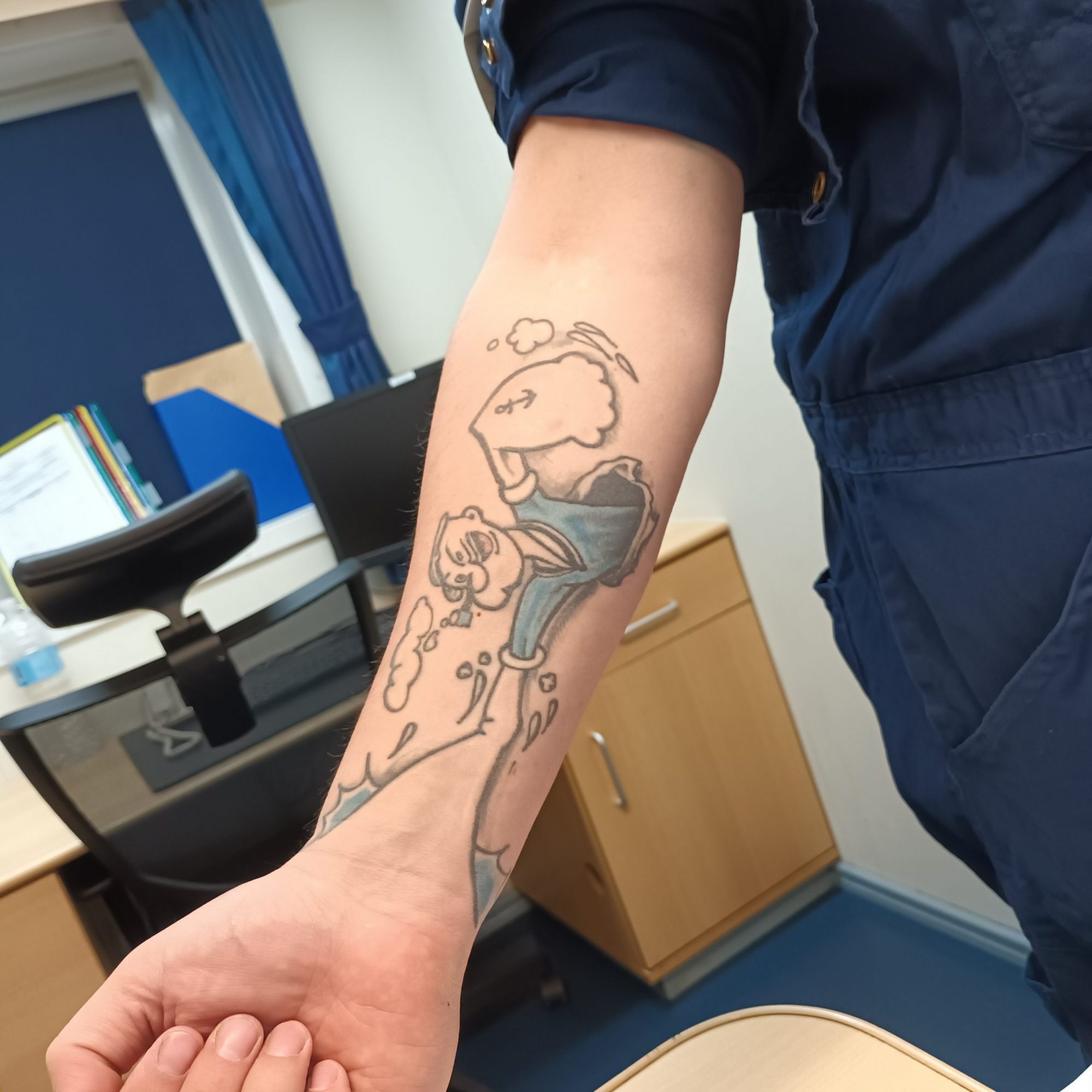 Zu sehen ist die Innenseite des rechten Unterarms eines Seemanns. Das Arbeitshemd ist hochgekrempelt, auf der Haut prangt ein Tattoo der Comicfigur Popeye.