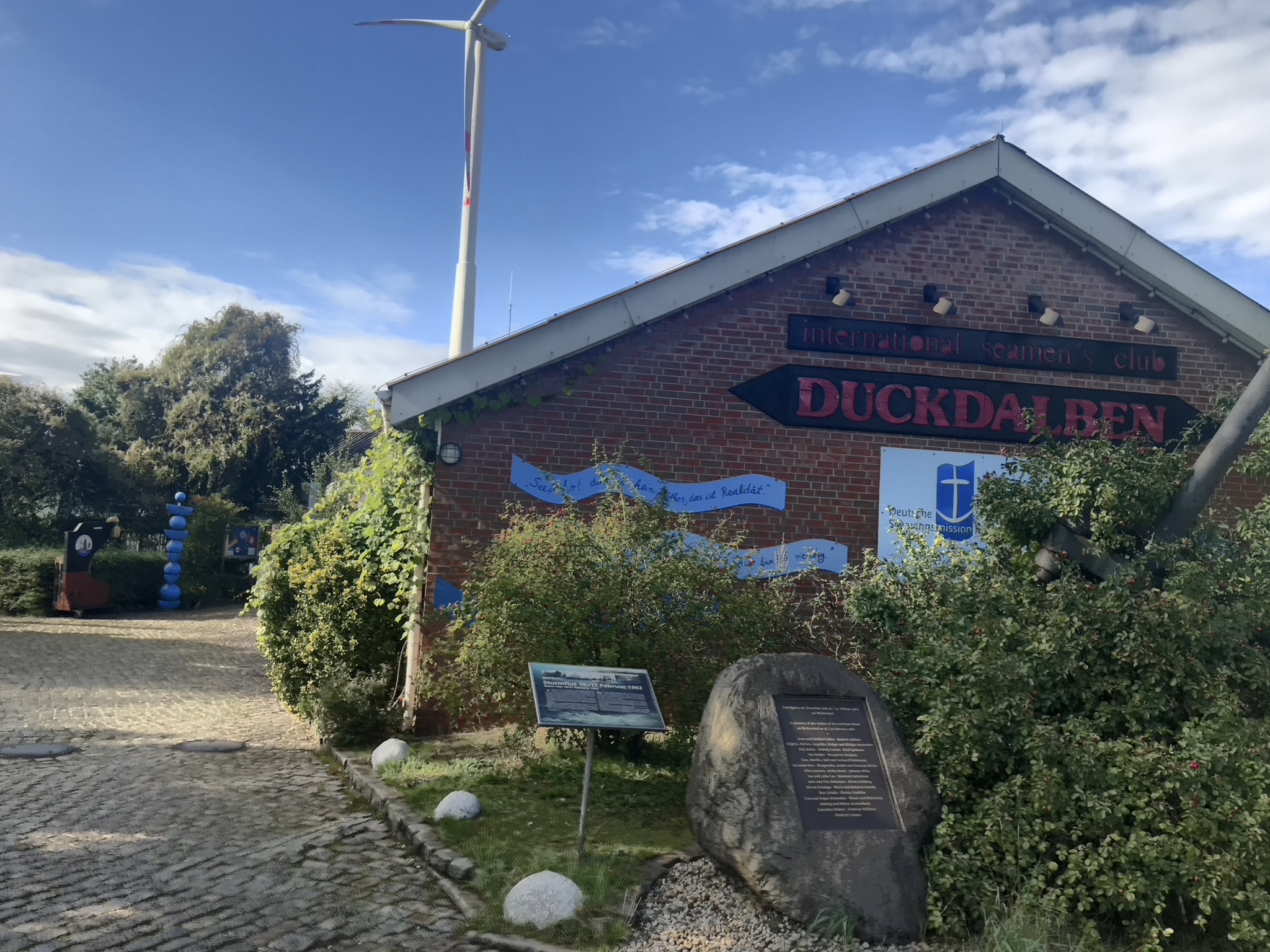 Blick auf die Seitenwand in roten Ziegeln mit dem Text international Seamen's club Duckdalben und dem Logo der Seemannsmission. Davor der Gedenkstein, ein grüner Busch und links davon der Weg mit Kopfsteinpflaster. Darüber der blaue Himmel und eine Windenergieanlage