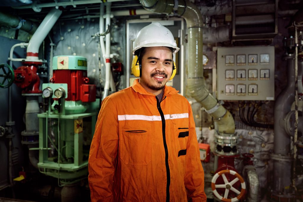 Das Foto zeigt einen Seemann, der in einem orangenen Arbeitsschutzanzeug gekleidet ist und einen weißen Schutzhelm und gelben Hörschutz trägt. Der Mann steht im Inneren eines Schiffes, wahrscheinlich im Maschinenraum, denn es sind Maschinen und eine Vielzahl an Rohren zu sehen. Foto: Joseph Heicks_Deutsche Seemannsmission