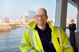 Matthias Ristau steht an die Reling gelehnt in gelber Warnjacke auf einem Schiff. Im Hintergrund ist die Elbphliharmonie zu sehen - DIe Lage der Seeleute