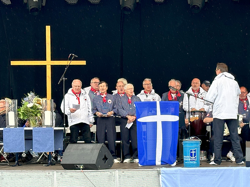 Das Foto zeigt eine Bühne, auf der der Shanty-Chor Mövenschiet singt. Links im Bild ist auch ein großes, schlichtes Kreuz aus hellem Holz zu sehen. DIe Sänger tragen blau-weiß gestreifte Seemannshemden und rote Halstücher. Einige der Männer tragen weiße Jacken. Gottesdienst zum Sea Sunday in Travemünde