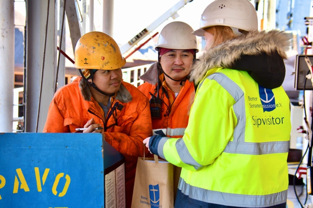 Das Foto zeigt eine ehrenamtliche Bordbesucherin der Bremer Seemannsmission, die von hinten zu sehen und an ihrer gelben Warnweste zu erkennen ist. Sie spricht an Bord eines Schiffes mit zwei asiatischen Seeleuten, die organge Arbeitsoveralls und Schutzhelme tragen. Die Bordbesucherin hält zwei Papiertüten in ihren Händen, die sie den Seeleuten übergibt.