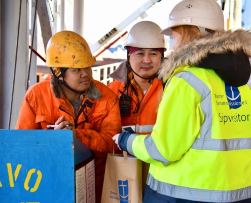 Das Foto zeigt eine ehrenamtliche Bordbesucherin der Bremer Seemannsmission, die von hinten zu sehen und an ihrer gelben Warnweste zu erkennen ist. Sie spricht an Bord eines Schiffes mit zwei asiatischen Seeleuten, die organge Arbeitsoveralls und Schutzhelme tragen. Die Bordbesucherin hält zwei Papiertüten in ihren Händen, die sie den Seeleuten übergibt.