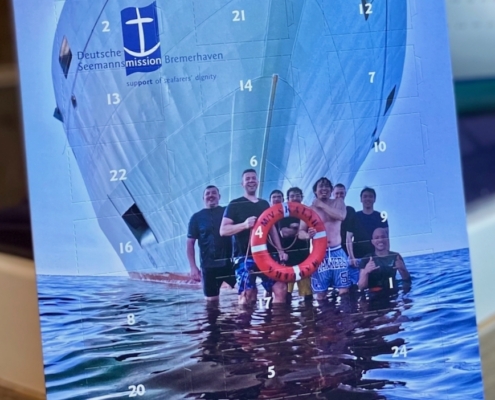 Maritimer Adventskalender 2025, man sieht den Bug eines Schiffes im Wasser, davor positioniert sind Seeleute, ein junger Mann in deren Mitte trägt einen Rettungsring um den Hals. Alle strahlen in die Kamera.