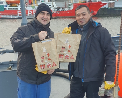 Das Foto zeigt zwei Seeleute an Bord eines Schiffes. Die beiden Männer halten Weihnachtstüten in den Händen und zeigen sie in die Kamera. Der Seemanns links im Bild trägt eine dunkle Mütze, beide tragen dunkle Arbeitskleidung. Der Seemannn rechts im Bild hat gelbe Arbeitshandschuhe an. Beide freuen sich über die Weihnachtsgeschenke der Seemannsmission Cuxhaven.