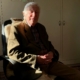 Das Foto zeigt den damals 96-jährigen Carl Osterwald, Pastor im Ruhestand und ehemaligen Generalsekretär der Deutschen Seemannsmission. Der ältere Herr mit schlohweißem Haar sitzt in einem olivfarbenen Wolljacket und mit rot-weiß kariertem Hemd auf einem schwarzem Bürodrehstuhl. Osterwald hat die Hände gefaltet und lächelt freundlich in die Kamera.