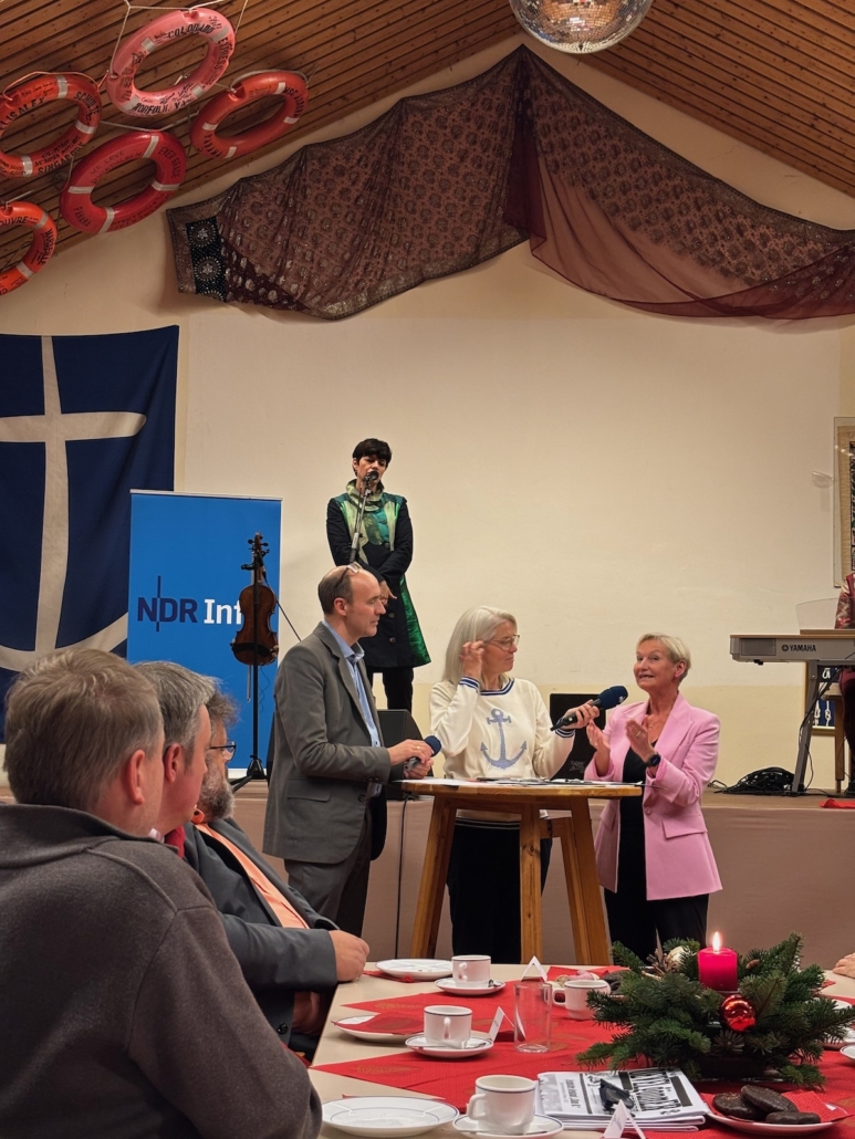Blick über einen weihnachtlich gedeckten Tisch, an dem Menschen sitzen hin zur Bühne, vor der an einem Stehtisch die Moderatorin und der Moderator mit Bischöfin Kirsten Fehrs stehen, die einen rosa Blazer trägt. Auf der Bühne steht eine Musikerin neben einer Geige vor dem Logo von NDR info und dem Anker Logo der Seemannsmission - Aufzeichnumg von Gruß an Bord im Duckdalben in Hamburg im Dezember 2024
