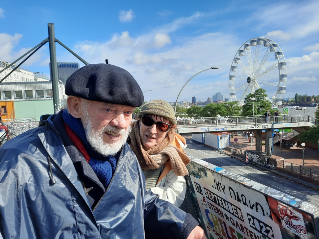Das Foto zeigt Hannes Schwartau (li.) und seine Frau Helga, die in der Nähe der Hamburger Landgunsbrücken auf die Elbe schauen. Im Hintergrund ist ein Riesenrad zu sehen, das an der Elbe steht. Die Sonne scheint, ein paar kleinere Wolken sind zu sehen, der Himmel ist blau. Beide tragen Mützen, Schals und Regenjacken.