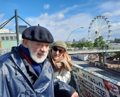 Das Foto zeigt Hannes Schwartau (li.) und seine Frau Helga, die in der Nähe der Hamburger Landgunsbrücken auf die Elbe schauen. Im Hintergrund ist ein Riesenrad zu sehen, das an der Elbe steht. Die Sonne scheint, ein paar kleinere Wolken sind zu sehen, der Himmel ist blau. Beide tragen Mützen, Schals und Regenjacken.