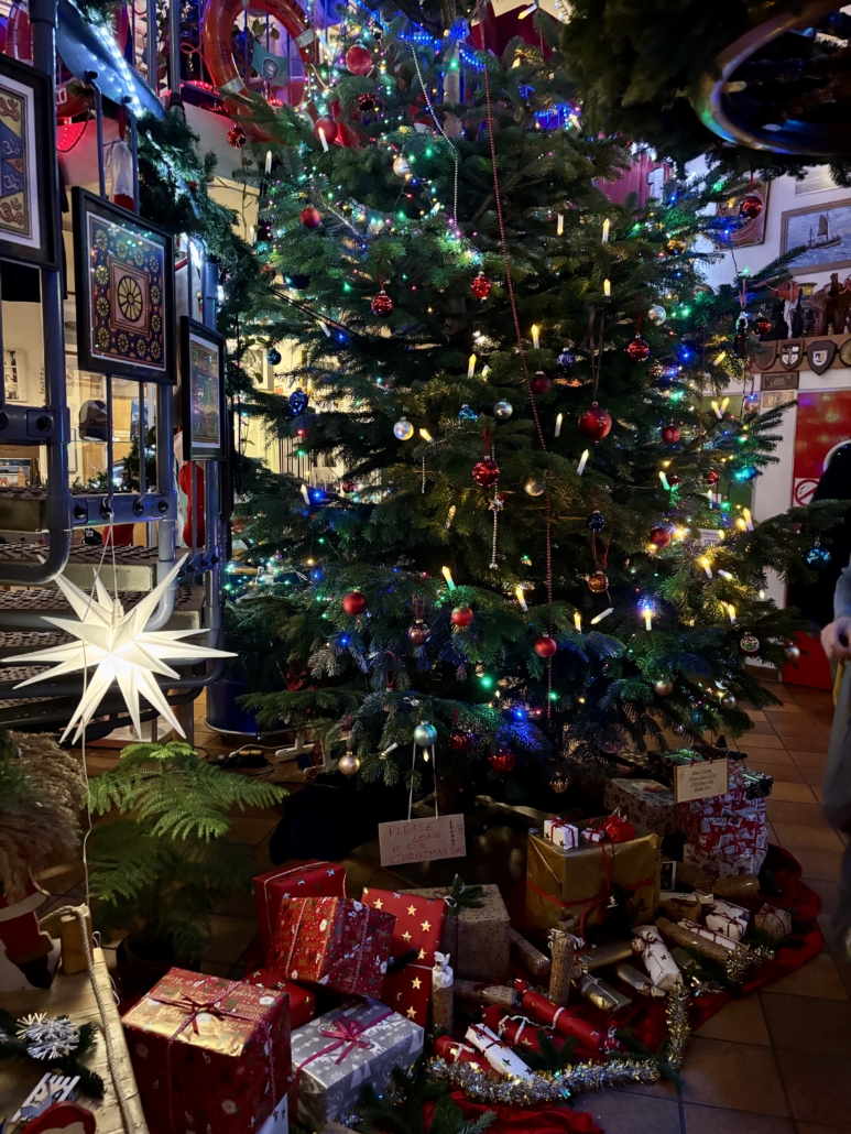 Unter dem großen Tannenbaum liegen viele schön verpackte Geschenke für Seeleute im Format von Schuhkartons für die Aktion Christmas in a Container Box
