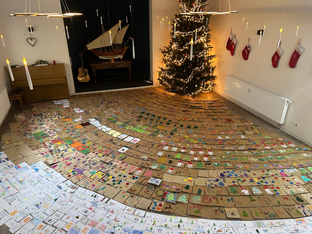Das Foto zeigt einen Raum, dessen Boden über und über mit individuell gestalteten Weihnachts-Papiertüten und -grußkarten bedeckt ist. Auf den Tüten und Karten sind Kindermalereien vom Schneemann, über weihnachtliche Zweige bishin zu bunten Kugeln zu sehen.