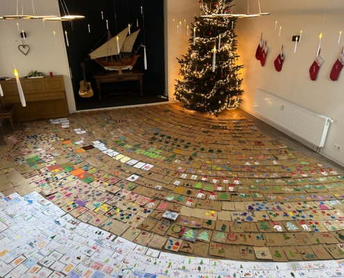 Das Foto zeigt einen Raum, dessen Boden über und über mit individuell gestalteten Weihnachts-Papiertüten und -grußkarten bedeckt ist. Auf den Tüten und Karten sind Kindermalereien vom Schneemann, über weihnachtliche Zweige bishin zu bunten Kugeln zu sehen.