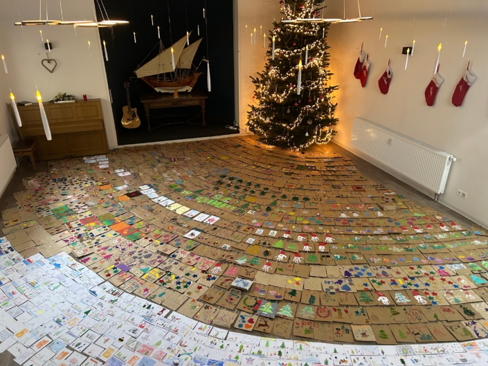 Das Foto zeigt einen Raum, dessen Boden über und über mit individuell gestalteten Weihnachts-Papiertüten und -grußkarten bedeckt ist. Auf den Tüten und Karten sind Kindermalereien vom Schneemann, über weihnachtliche Zweige bishin zu bunten Kugeln zu sehen.