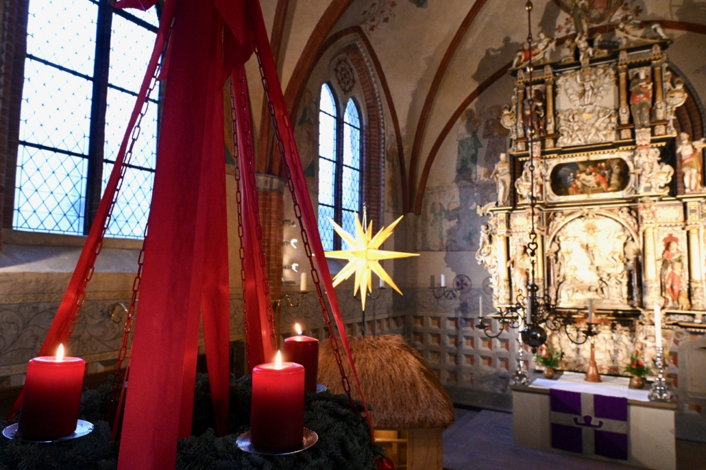 Das Foto zeigt großen Adventskranz, der an roten Ketten hängt, mit roten Seidenbändern verziert ist und drei brennende Kerzen trägt. Unscharf im Hintergrund sieht man einen mittelalterlichen Dorfkirchenaltar.