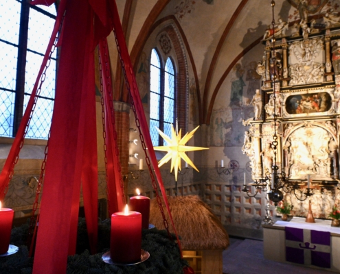 Das Foto zeigt großen Adventskranz, der an roten Ketten hängt, mit roten Seidenbändern verziert ist und drei brennende Kerzen trägt. Unscharf im Hintergrund sieht man einen mittelalterlichen Dorfkirchenaltar.