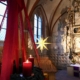Das Foto zeigt großen Adventskranz, der an roten Ketten hängt, mit roten Seidenbändern verziert ist und drei brennende Kerzen trägt. Unscharf im Hintergrund sieht man einen mittelalterlichen Dorfkirchenaltar.