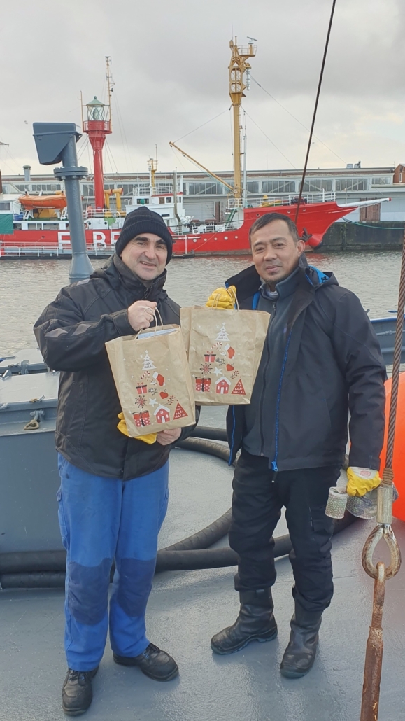 Das Foto zeigt zwei Seeleute an Bord eines Schiffes. Die beiden Männer halten Weihnachtstüten in den Händen und zeigen sie in die Kamera. Der Seemanns links im Bild trägt eine dunkle Mütze, beide tragen dunkle Arbeitskleidung. Der Seemannn rechts im Bild hat gelbe Arbeitshandschuhe an. Beide freuen sich über die Weihnachtsgeschenke der Seemannsmission Cuxhaven.