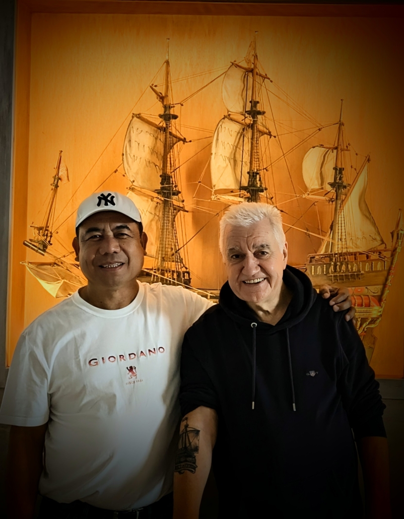 Das Foto zeigt zwei Männer. Rechts im Bild ist der Ehrenamtliche Peter Dany von der Deutschen Seemannsmission in Altona zu sehen. Er trägt einen schwarzfarbenen Hoodie und hat seinen rechten Ärmel hochgekrempelt, sodass ein Tatoo zu sehen ist. Es zeigt ein Segelschiff. Der Herr links im Bild trägt ein weißes T-Shirt und eine weiße Baseballkappe. Er hat seinen Arm um Peter Danys Schulter gelegt. Beide lächeln freundlich in die Kamera.
