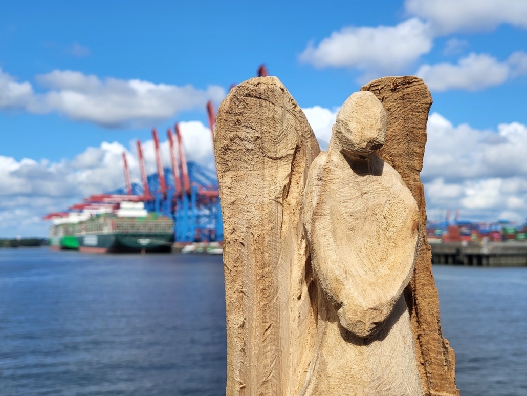 Das Foto zeigt einen grob geschnitzten Holzengel, der die Hände wie zum Gebet gefaltet hat. Die Figur ist aus hellem Holz geschnitzt. Im Hintergrund sieht man die Silhouette des Hamburger Hafens. Es ist ein sonniger Tag, ein paar Wolken ziehen über den ansonsten blauen Himmel. Im Hintergrund sieht man unscharf die blau-rotgestrichenen Hafenkräne und ein Containerschiff, dessen bunte Container gerade be- oder entladen werden.