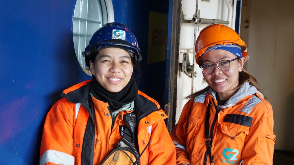 Zwei Frauen in Arbeitsbekleidung und mit Schutzhelmen lehnen an einer dunkelblau gestrichenen Tür, wahrscheinlich an Bord eines Schiffes. Ihre Arbeitsoveralls und Schutzjacken sind orange mit Relektorstreifen. Beide Frauen lächeln freundlich in die Kamera und kommen wahrscheinlich aus Asien. Die Frau auf der linken Seite des Fotos trägt ein schwarzes Funkgerät.