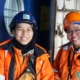 Zwei Frauen in Arbeitsbekleidung und mit Schutzhelmen lehnen an einer dunkelblau gestrichenen Tür, wahrscheinlich an Bord eines Schiffes. Ihre Arbeitsoveralls und Schutzjacken sind orange mit Relektorstreifen. Beide Frauen lächeln freundlich in die Kamera und kommen wahrscheinlich aus Asien. Die Frau auf der linken Seite des Fotos trägt ein schwarzes Funkgerät.