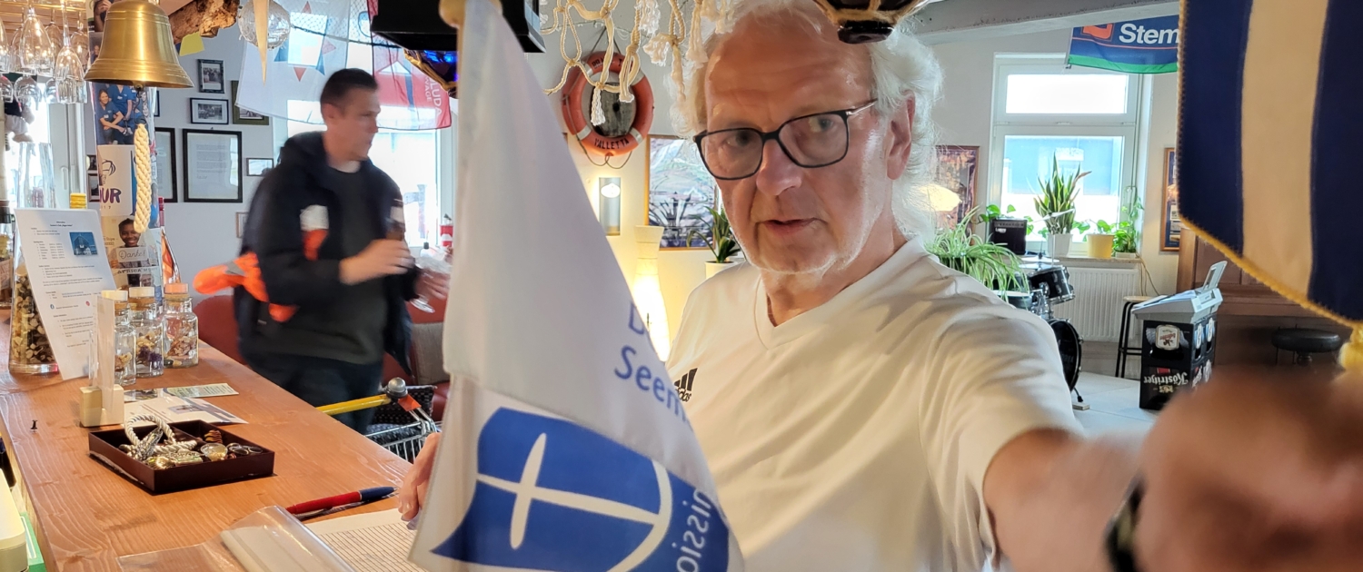 Peter Leukroth im Seemannsclub Rügenanker schaut über die Bar hinweg. Er trägt ein schlichtes weißes T-Short, reine schwarzumrandete Brille und hat schlohweißes, lockiges Haar.
