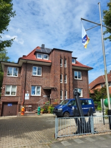 Das Foto zeigt das Seemannsheim der Deutschen Seemannsmission in Wilhelmshaven. Das Haus ist eine Backsteinvilla aus der Zeit um die Jahrhundertwende. Rechts vor dem Haus gibt es Parkplätze. Von der Straßenseite aus links steht ein großer Baum. Hinter dem Haus befindet sich ein Garten, in dem die Seeleute in den wärmeren Monaten entspannen können.