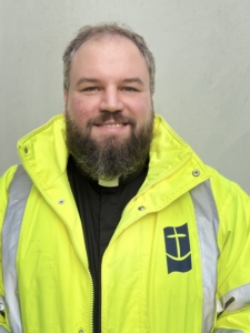 Das Foto zeigt André Schuller, der die Deutsche Seemannsmission in Rotterdam leitet. Er trägt eine neon-gelbe Arbeitsjacke mit dem Logo der Deutschen Seemannsmission, dem sogenannten lachenden Anker auf marineblauem Hintergrund. DIe Jacke hat zwei Reflektionsstreifen. Der 34-jährige Hamburger trägt einen Bart.