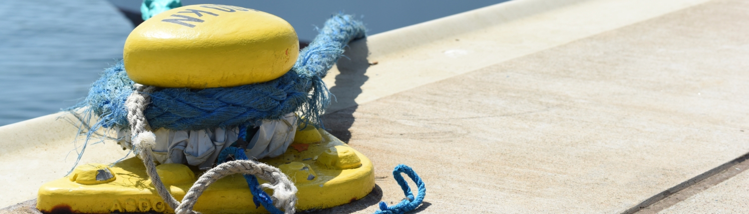 Auf diesem Symbolbild ist ein leuchtend gelb angestrichener Poller an einer Kaimauer im Hafen zu sehen. Er ist fest im Boden verankert und dient dazu, Schiffe sicher an der Kaimauer festzumachen. An einem blauen Tau sind unscharf im Hintergrund Teiles eines Schiffsbugs (vorn) zu erkennen.