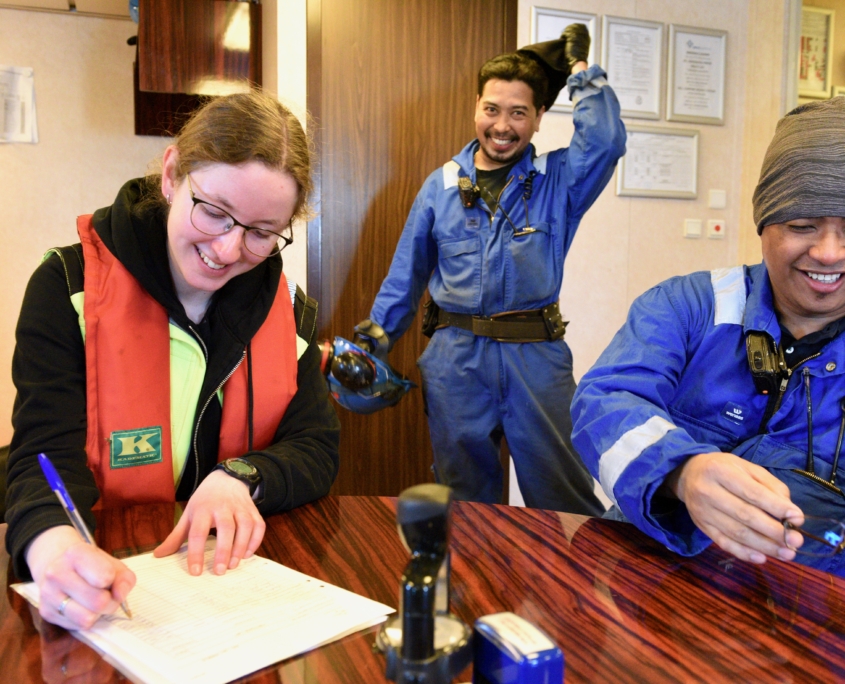 Das Foto zeigt eine FSJlerin der Deutschen Seemannsmission Brunsbüttel. Sie füllt ein Formular aus, wahrscheinlich ein Dokument, um eine Telefonkarte zu registrieren. Neben ihr am Tisch sitzt ein Seemann in einem blauben Arbeitsoverall. Im Hintergrund steht ein Seemann, der sich die Mütze vom Kopf zieht und lächelnd in die Kamer schaut.