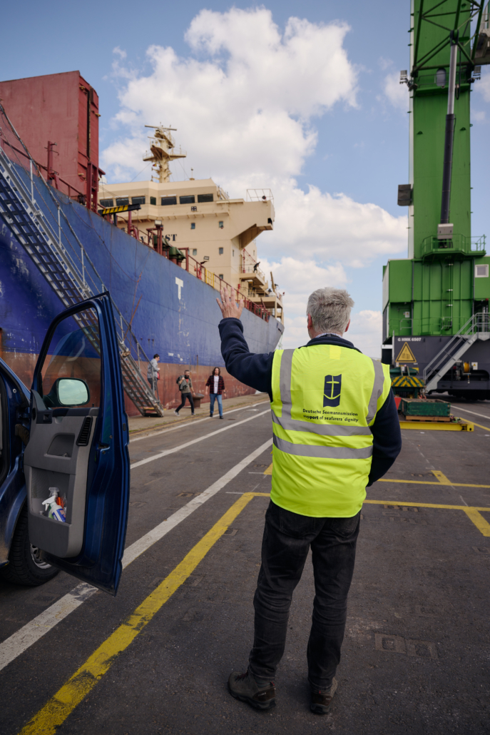 Das Foto zeigt Seemannspastor Marc Schippers im Hafen von Antwerpen. Er ist nur von hinten zu sehen und trägt eine Sicherheitswester mit dem blauben Logo der Deutschen Seemannsmission. Er ist gerade aus seinem Dienstwagen gestiegen und steht an der Kaikante in der nähe eines Containerschiffs. Der Himmel ist blau, mit ein paar kräftigen weißen Wolken.