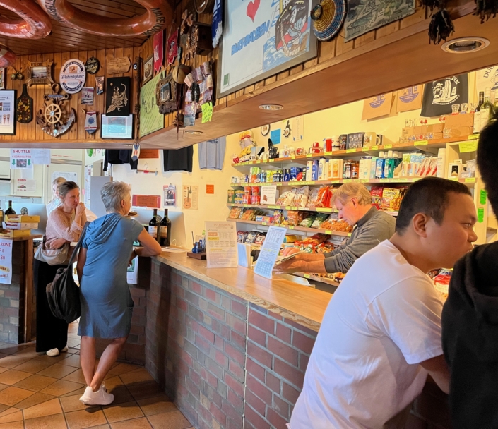 Das Foto zeigt die Theke im Seemannsclub Duckdalben der Deutschen Seemannsmission Hamburg-Harburg. Man sieht einen kleinen Laden, in dem die Seeleute waren wie Hygieneartikel, Süßigkeiten oder Chips kaufen können. Auch Telefonkarten können gekauft und Geld gewechselt werden. An der Decke hängen zahlreiche Rettungsringe von Schiffen.