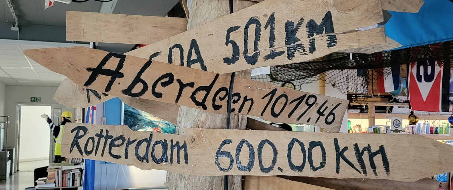 Holz-Wegweiser mit Entfernungsmarken in alle Himmelsrichtungen, z. B. Rio oder Aberdeen. Der Wegweiser steht im Seemannsclub Rügen Anker im Fährhafen von Mukran auf Rügen.