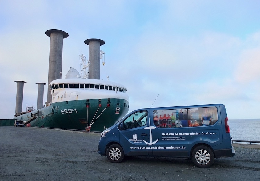 Das Foto ist im Hafen von Cuxhaven entstanden. Es zeigt ein Schiff mit Aufbauten, das an der Kaikante festgemacht ist. Davor steht leicht schräg der Bulli der Seemannsmission Cuxhaven, mit dem die Seeleuten von den Schiffen abgeholt und in den Seemannsclub gebracht werden.