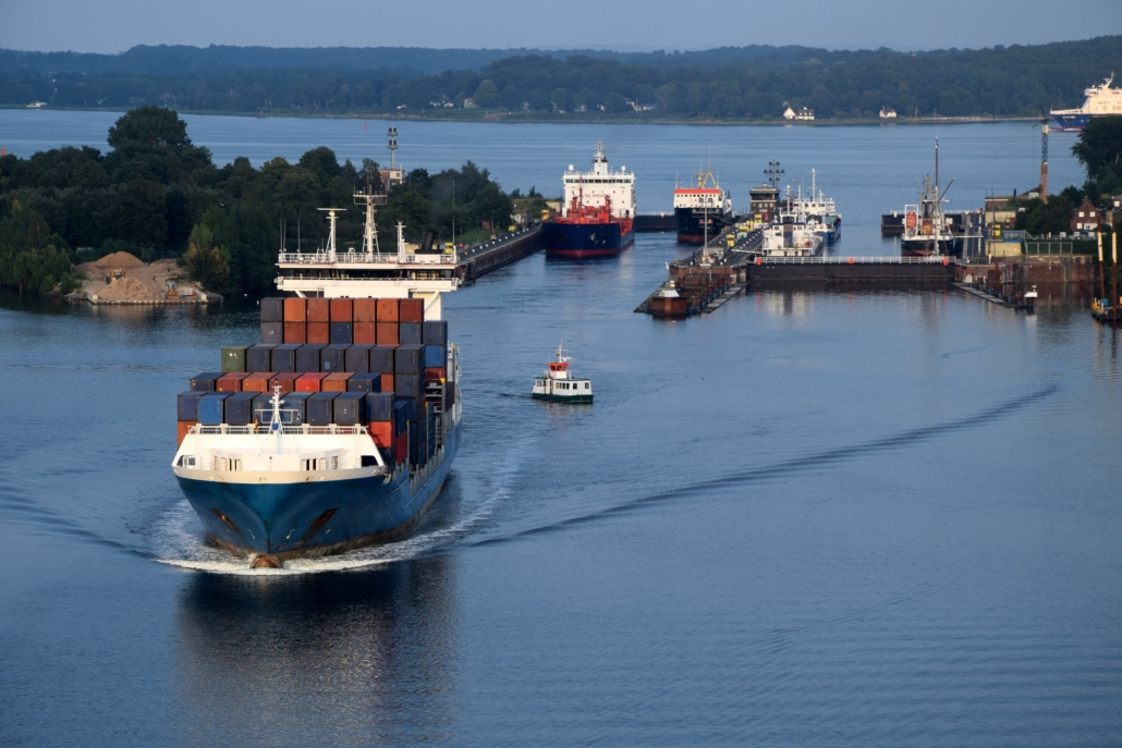Das Foto zeigt die Schleusen in Kiel-Holtenau des Nord-Ostsee-Kanals. Vorn links im Bild sieht man ein Containerschiff mit vielen Containern in Rot-, Orange-, Grün- und Blautönen. Die Wasseroberfläche ist ruhig, zu sehen ist eine Abend- oder Morgenstimmung.