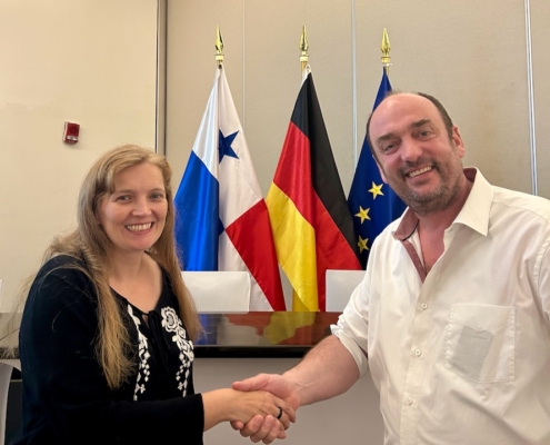 Andrea Meenken (links), Stationsleiterin der Deutschen Seemannsmission Panaman reicht Magnus Deppe die Hand. Deppe leitet die Bremer Seemannsmission. Hinter den beiden sind die Flagge von Panama, die deutsche udn die der Europäischen Union zu sehen. Andrea Meenken trägt ihr langes, blondes Haar offen und ein schwarzes, langes Oberteil. Deppe trägt ein weißes, kurzärmeliges Businesshemd.