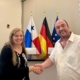 Andrea Meenken (links), Stationsleiterin der Deutschen Seemannsmission Panaman reicht Magnus Deppe die Hand. Deppe leitet die Bremer Seemannsmission. Hinter den beiden sind die Flagge von Panama, die deutsche udn die der Europäischen Union zu sehen. Andrea Meenken trägt ihr langes, blondes Haar offen und ein schwarzes, langes Oberteil. Deppe trägt ein weißes, kurzärmeliges Businesshemd.
